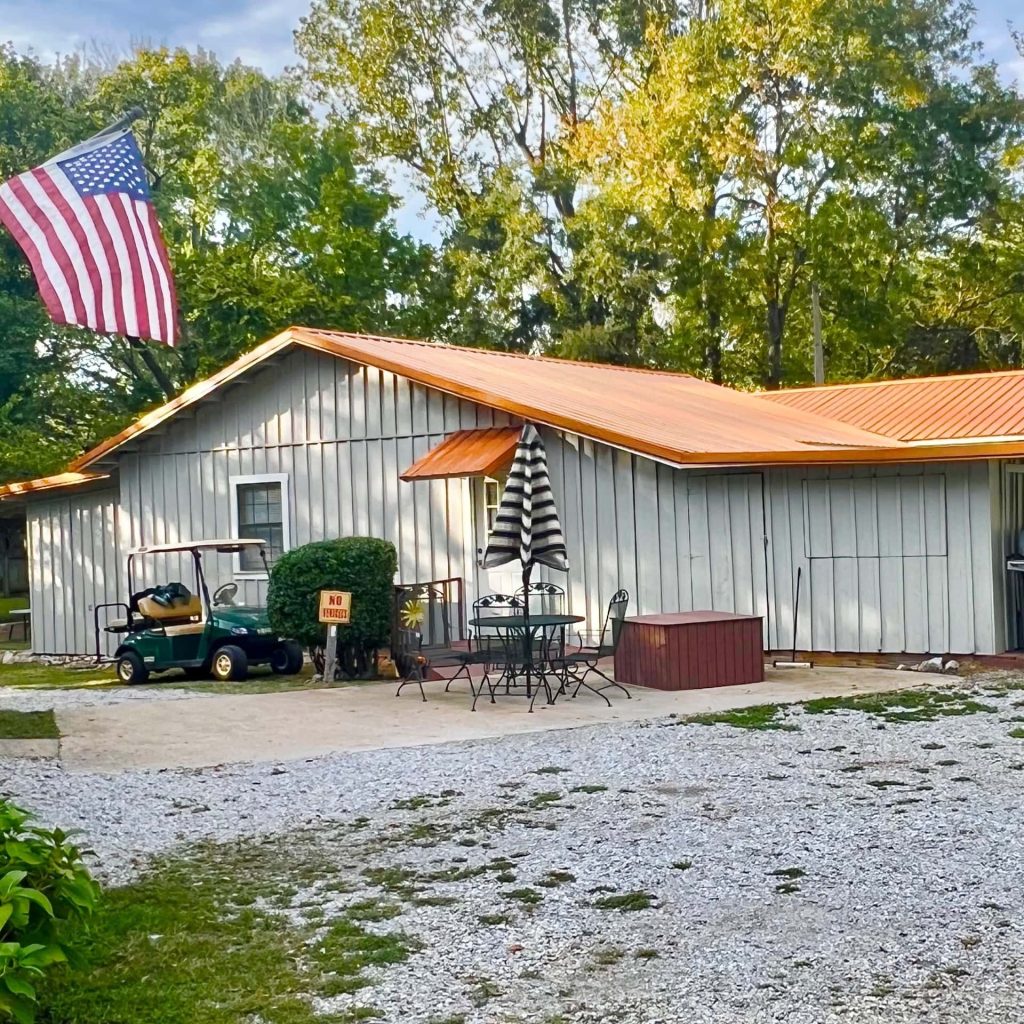 A white building with a yellow roof, an American flag, a golf cart, patio furniture, and trees in the background. Gravel driveway in the foreground.