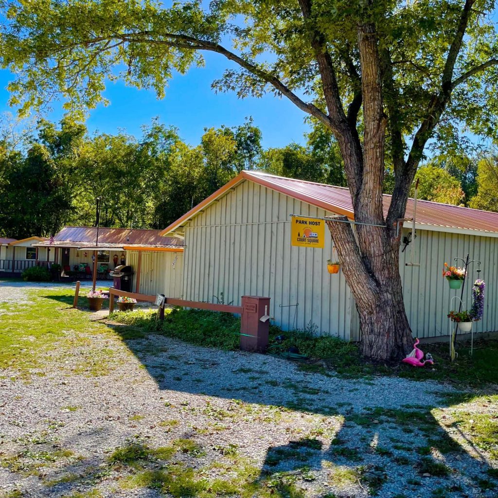 A rustic building with a corrugated metal roof, surrounded by trees, features a small parking area with gravel ground and various hanging plants and decorations on the exterior.