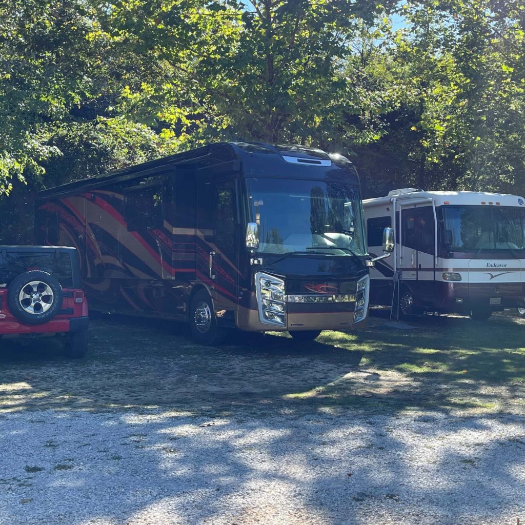 Two RVs and a red Jeep are parked on a gravel area surrounded by trees.