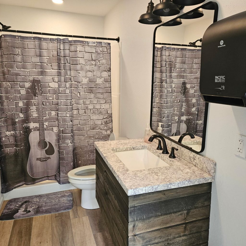 A bathroom with wood accents, a rectangular sink, black fixtures, a large paper towel dispenser, and a shower curtain featuring a guitar against a brick wall.