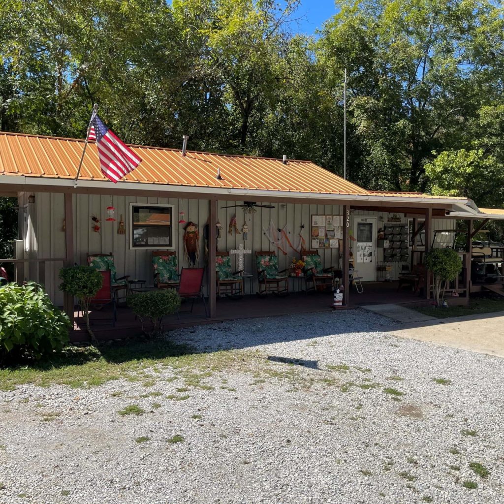 A small, rustic building with a metal roof and an American flag stands in a wooded area. The porch has chairs and decorations. A gravel area is in the foreground.