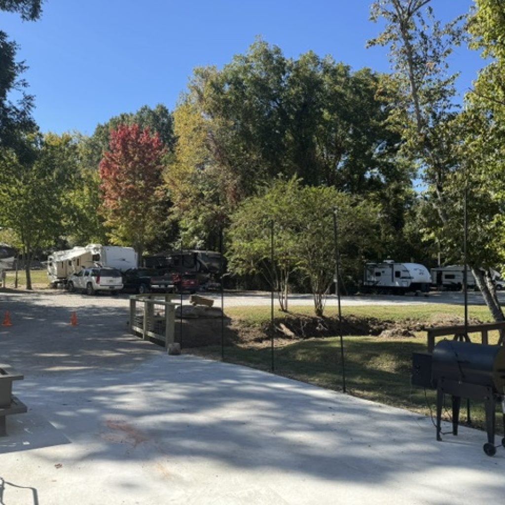 Several RVs parked in a wooded campground with a concrete patio and grill in the foreground.