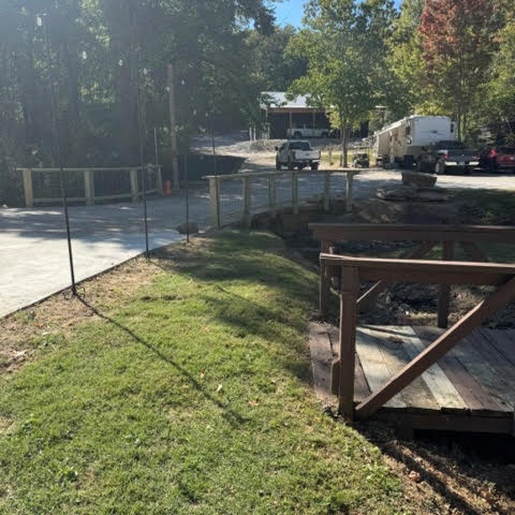 A wooden bridge crosses a small ditch near a gravel driveway with parked vehicles and a building in the background.