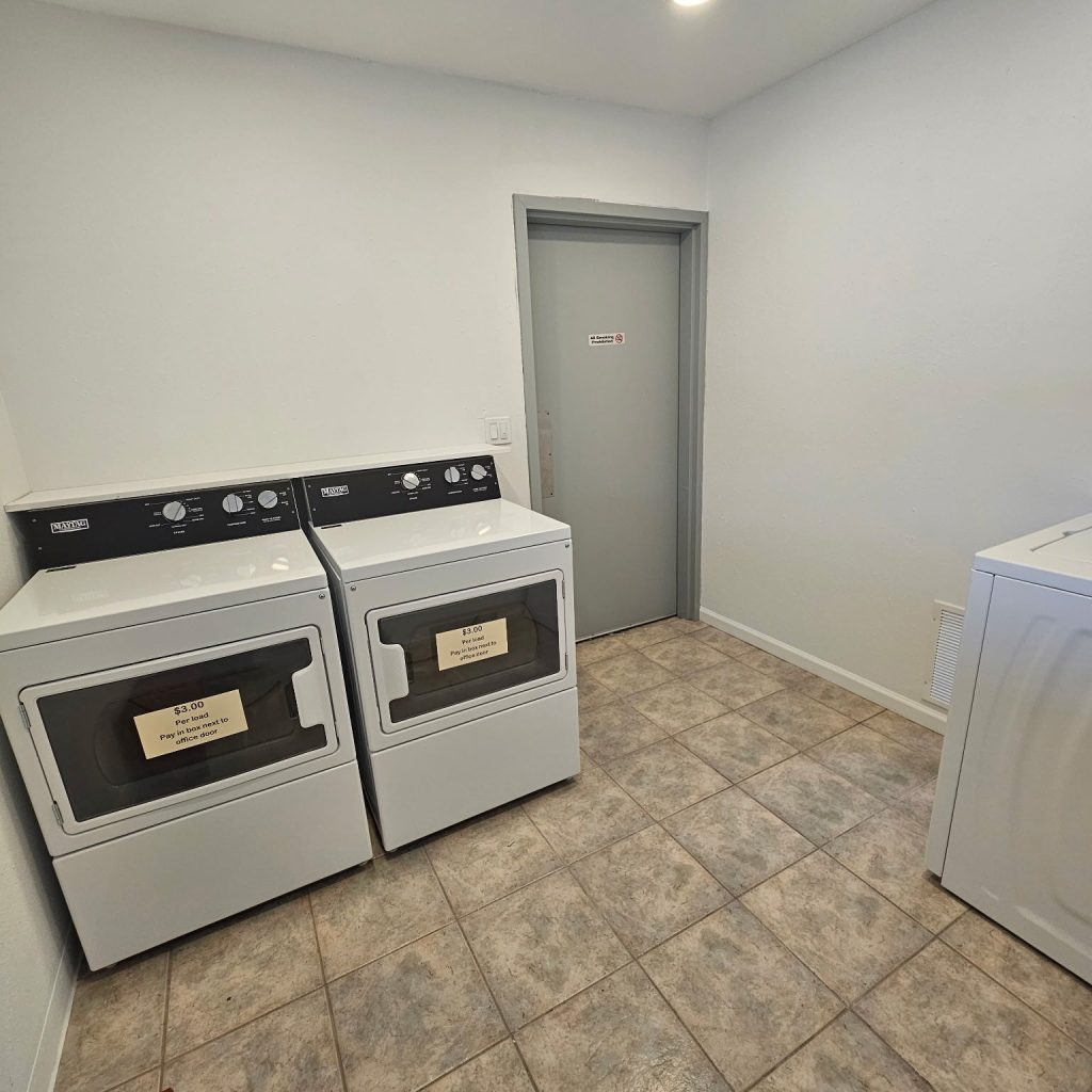 A laundry room with two dryers side by side, a washing machine on the right, tiled floor, white walls, and a closed gray door in the background.